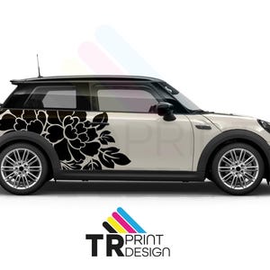 Può includere: Una Mini Cooper bianca con una decalcomania floreale nera sul lato. La decalcomania presenta un grande fiore e foglie. L'auto ha passaruota neri e un tetto nero. Le ruote sono argentate. Il logo "TR PRINT DESIGN" è in basso.