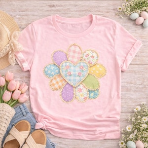 May include: A light pink t-shirt featuring a colorful floral design. The design includes a heart-shaped center and petal-like shapes in various pastel patterns. The shirt has short sleeves and a crew neck.