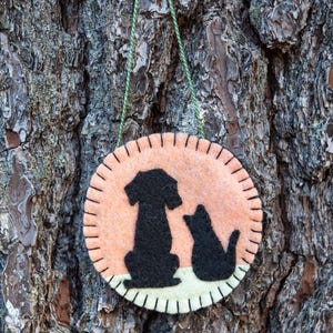 May include: A round felt ornament featuring a black silhouette of a dog and a cat. The ornament has a peach-colored center with black stitching around the edge. A green string is attached for hanging.