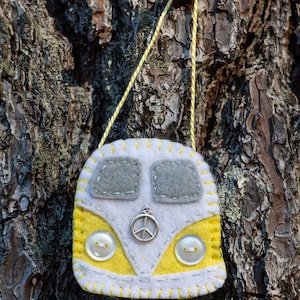 May include: A felt ornament shaped like a yellow and white camper van with gray accents. The ornament features a peace sign charm, two white buttons, and yellow stitching. A yellow string is attached for hanging.