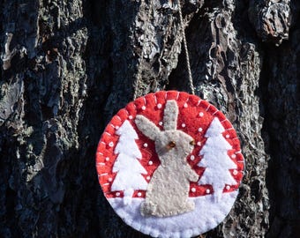 May include: A round, felt Christmas ornament with a red background and a white border. The ornament features a light gray bunny and two white Christmas trees. White dots are scattered across the red background, and the ornament hangs from a string.