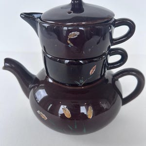 May include: A stackable dark brown ceramic tea set consisting of a teapot, a creamer, and a lidded sugar bowl. Each piece features a glossy finish and is decorated with small, hand-painted wheat stalks in shades of gold, white, and green.