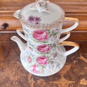 May include: A stackable porcelain tea set for one, featuring a white base with pink rose floral patterns, green leaves, and gold trim. The set includes a teapot, a teacup, and a lid topped with a sculpted white rose flower handle.