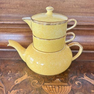 May include: A yellow ceramic tea set for one, featuring a stackable design with a teapot base, a middle cup, and a top lid. The pieces have a speckled yellow finish, gold-toned rims, and a gold-toned handle on the teapot and the cup.