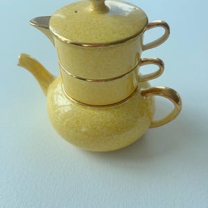 May include: A yellow tea set for one, featuring a teapot base with a stacked cup and lid. The ceramic pieces have a speckled yellow finish with gold-toned trim on the handles, rims, and the lid knob. The design allows the cup to nest on the teapot.