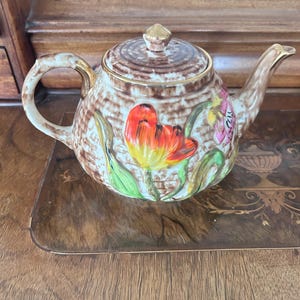 Peut inclure: Une théière en céramique avec un motif tressé marron texturé, ornée de tulipes rouges et jaunes en relief peintes à la main et de feuilles vertes. Elle comprend un couvercle assorti avec un bouton doré, une anse courbée et un bec verseur doré.