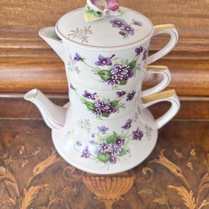 Könnte beinhalten: Ein weißes Porzellan-Teeservice für eine Person, vertikal gestapelt, mit handgemalten violetten Veilchen und grünen Blättern. Das Set besteht aus einer Teekanne, einer Teetasse und einem Deckel mit einer skulpturalen, dreidimensionalen Blume.