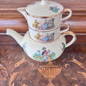 May include: A cream-coloured ceramic tea set for one, featuring a stacked teapot and cup with gold trim. The pieces are decorated with colourful illustrations of 18th-century figures, including an artist painting a portrait and outdoor scenes.
