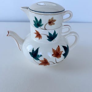 May include: A cream-coloured ceramic tea set for one, consisting of a teapot base and a stacked cup with a lid. The pieces feature a hand-painted pattern of green and orange maple-style leaves with thin brown stems on a smooth, glossy surface.