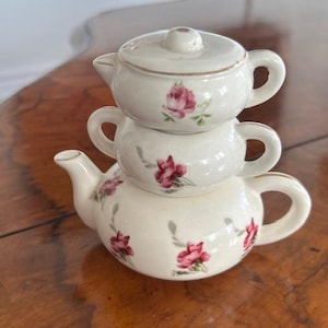 Può includere: Servizio da tè in ceramica color crema impilato con una teiera, una zuccheriera e una lattiera. Ogni pezzo è adornato con motivi floreali di rose rosa e finiture dorate. La teiera ha un beccuccio e un manico.