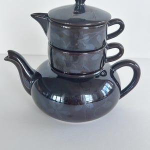 May include: A dark brown ceramic tea set for one, featuring a stackable design with a teapot at the base, a cup in the middle, and a lidded creamer or sugar bowl on top. The glossy surface is decorated with a subtle, tonal floral pattern.