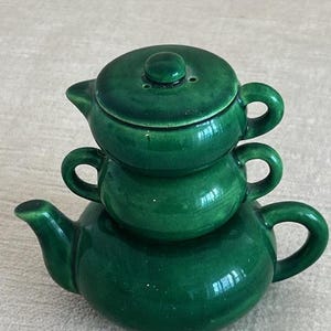 Può includere: Una pila di tre teiere in ceramica verde. La teiera inferiore ha un beccuccio e un manico, quella centrale ha un manico e quella superiore ha un coperchio con un piccolo pomello. Le teiere sono impilate l'una sull'altra.