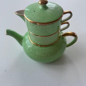 May include: A stacked ceramic tea set featuring a light green speckled glaze with gold trim. The set consists of a rounded teapot base with a curved spout and handle, topped by a matching cup with a lid, both accented with gold bands.