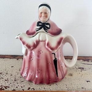 May include: A ceramic teapot shaped like a person wearing a pink dress with a white collar and a black bonnet. The teapot features a black bow at the neck, a black umbrella graphic on the skirt, and small black dots scattered across the pink fabric.