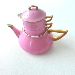 May include: A pink ceramic tea set for one, featuring a teapot base with a gold-trimmed handle and spout, topped with a matching stacked cup and lid. The set is finished with gold accents on the lid handle, cup rim, and cup handle, creating a cohesive look.