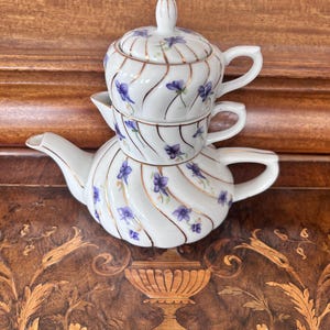 May include: A white porcelain tea set for one, stacked vertically, featuring a teapot, creamer, and lidded sugar bowl. Each piece has a swirling ribbed texture, gold-painted trim, and a repeating pattern of small purple violet flowers.