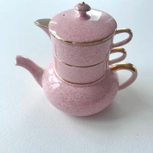 May include: A stackable tea set for one featuring a pink speckled ceramic finish with gold trim. The set includes a teapot base with a curved spout and handle, topped by a matching cup with a handle and a lid with a small gold-toned knob.