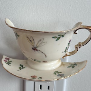 Könnte beinhalten: Ein cremefarbenes Keramik-Nachtlicht für die Steckdose in Form einer Teetasse mit Untertasse. Das Stück zeigt eine handgemalte Libelle, kleine grüne Blätter und bunte Blumenmotive. Der Rand der Tasse und der Untertasse ist goldfarben verziert.