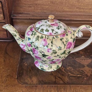 Könnte beinhalten: Eine Keramik-Teekanne mit hellgelbem Hintergrund und einem Muster aus rosa Rosen und grünen Efeublättern. Die Teekanne hat einen geschwungenen Griff, einen langen Ausguss und einen passenden Deckel mit einem goldfarbenen Knauf. Rand und Griff sind goldfarben.