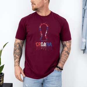 A man wearing a burgundy t-shirt with a graphic of a trophy and the word "Croatia" printed on it, standing against a white wall.