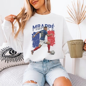 A woman wearing a white sweatshirt with a graphic of a soccer player and the text "MBARPE" is sitting on a bed, holding a coffee mug.