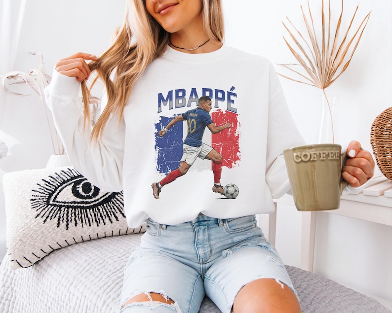 A woman wearing a white sweatshirt with a graphic of a soccer player and the text "MBARPE" is sitting on a bed, holding a coffee mug.