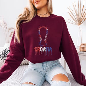 A woman wearing a burgundy sweatshirt with a graphic of a trophy and the word "Croatia" printed on it, sitting on a bed.