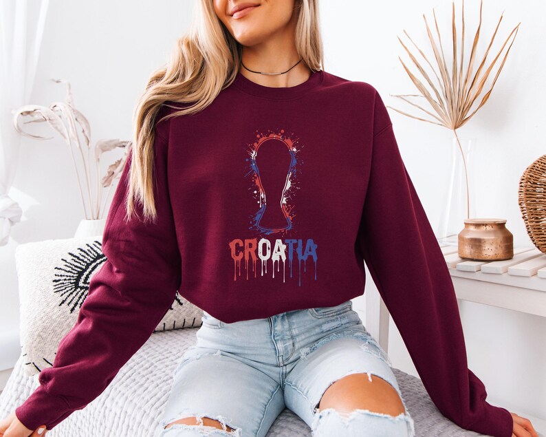 A woman wearing a burgundy sweatshirt with a graphic of a trophy and the word "Croatia" printed on it, sitting on a bed.