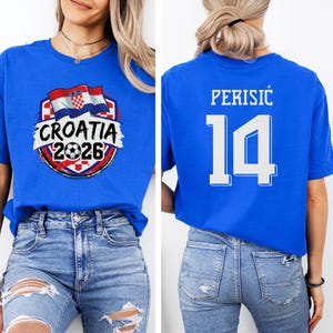 A woman wearing a blue t-shirt with a graphic of the Croatia national flag and the text "CROATIA 2026" printed on it. The t-shirt also has the number 14 printed on the back.
