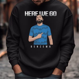 A man wearing a black sweatshirt with a graphic design of a man pointing and the text "HERE WE GO BENZEMA" printed on it.