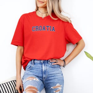 A woman wearing a red t-shirt with the word "Croatia" printed on it, paired with distressed blue jeans, stands in front of a white wall.