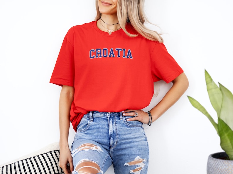 A woman wearing a red t-shirt with the word "Croatia" printed on it, paired with distressed blue jeans, stands in front of a white wall.