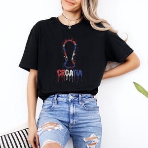 A woman wearing a black t-shirt with a graphic design and jeans stands against a white wall, with a potted plant visible in the background.