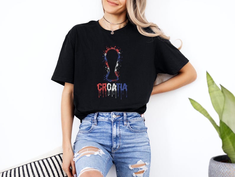 A woman wearing a black t-shirt with a graphic design and jeans stands against a white wall, with a potted plant visible in the background.