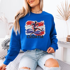 A woman wearing a blue sweatshirt with a graphic of a soccer player and the word "Croatia" printed on it, sitting on a couch.