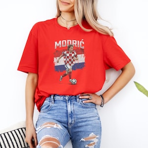 A woman wearing a red t-shirt with a graphic of a soccer player and jeans stands in front of a white wall.