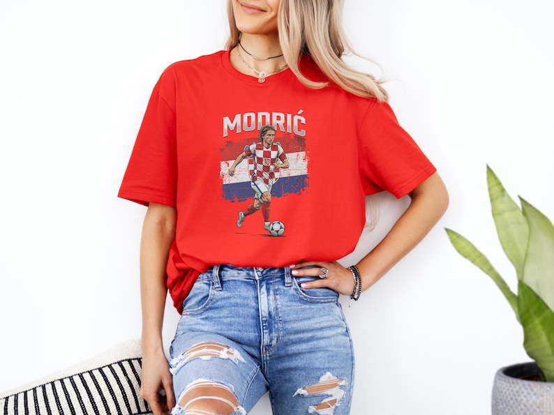 A woman wearing a red t-shirt with a graphic of a soccer player and jeans stands in front of a white wall.