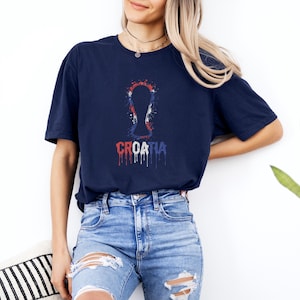 A woman wearing a navy blue t-shirt with a graphic design and jeans stands against a white wall, with a potted plant visible in the background.