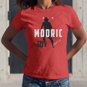 A red t-shirt with a graphic design featuring a silhouette of a person and the text "MODRIC" printed on it.