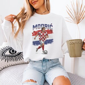 A woman wearing a white sweatshirt with a graphic of a soccer player is sitting on a bed, holding a coffee mug.