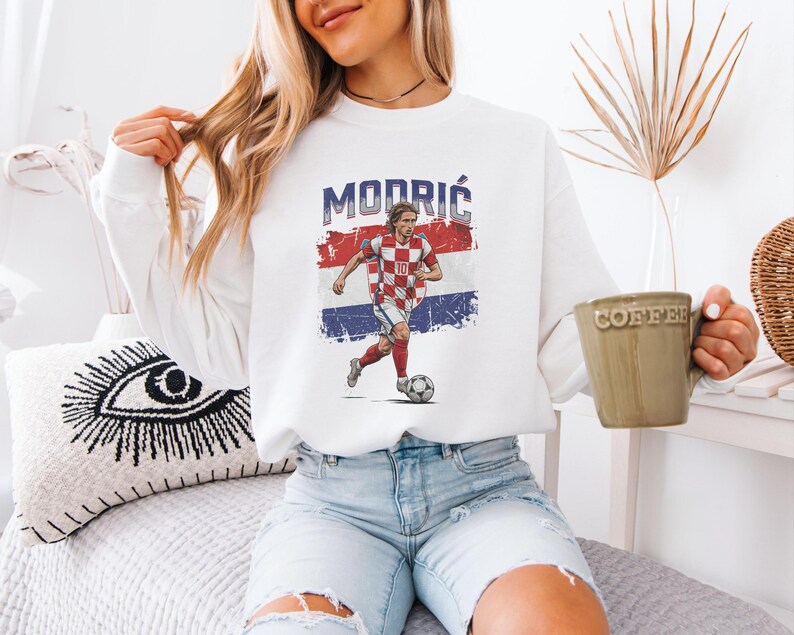 A woman wearing a white sweatshirt with a graphic of a soccer player is sitting on a bed, holding a coffee mug.