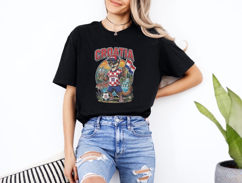 A woman wearing a black t-shirt with a graphic of a bear holding a flag and a soccer ball, standing against a white wall.