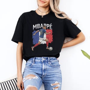 A woman wearing a black t-shirt with a graphic of a soccer player and the text "MBARPE" is standing in front of a white wall.