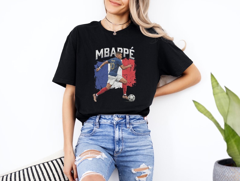 A woman wearing a black t-shirt with a graphic of a soccer player and the text "MBARPE" is standing in front of a white wall.