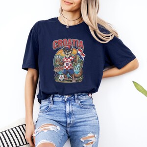 A woman wearing a navy blue t-shirt with a graphic of a cartoon character and the word "Croatia" printed on it, standing against a white wall.