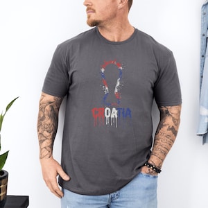 A man wearing a gray t-shirt with a graphic design and text printed on it, standing next to a plant.