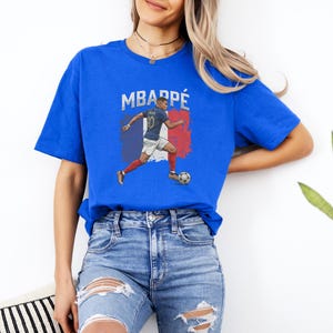A woman wearing a blue t-shirt with a graphic of a soccer player and jeans, standing against a white wall.