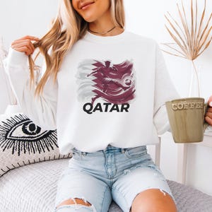 A woman wearing a white sweatshirt with a graphic of a bird and the word "QATAR" is sitting on a bed, holding a coffee mug.
