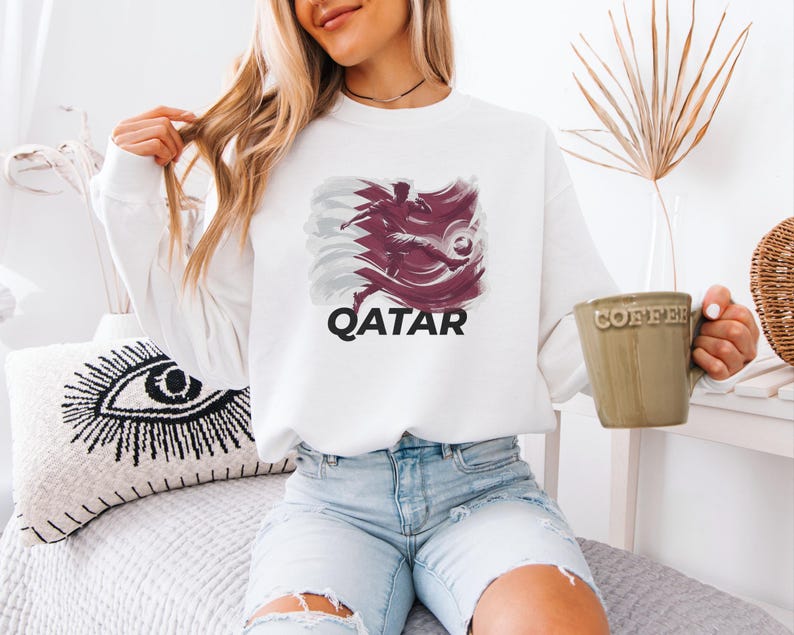 A woman wearing a white sweatshirt with a graphic of a bird and the word "QATAR" is sitting on a bed, holding a coffee mug.
