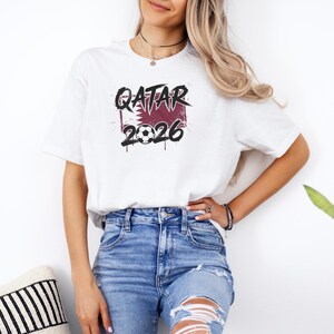 A woman wearing a white t-shirt with the text "Qatar 2016" printed on it, standing next to a potted plant.